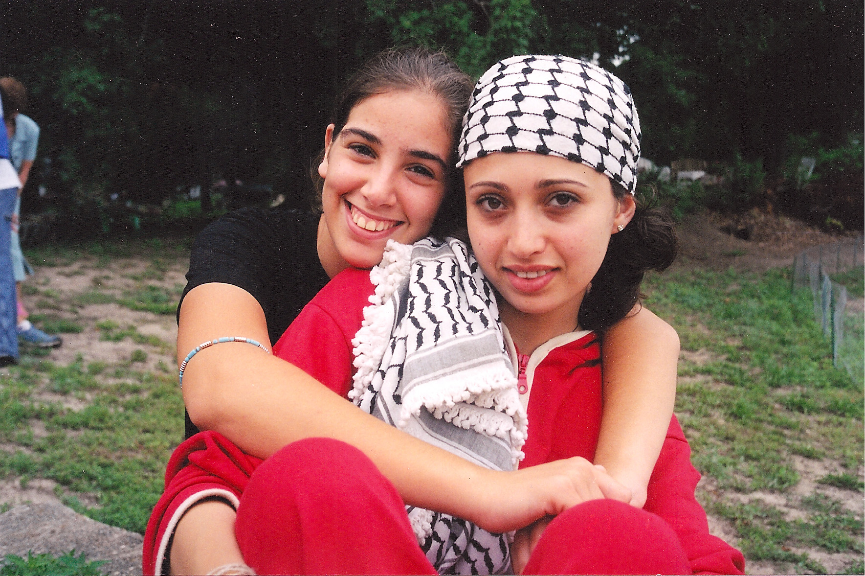 A Palesintian teen girl with a keffiyeh around her head sits on the grass. Behind her, and with arms wrapped around her, is an Israeli teen girl. They both smile for the camera.